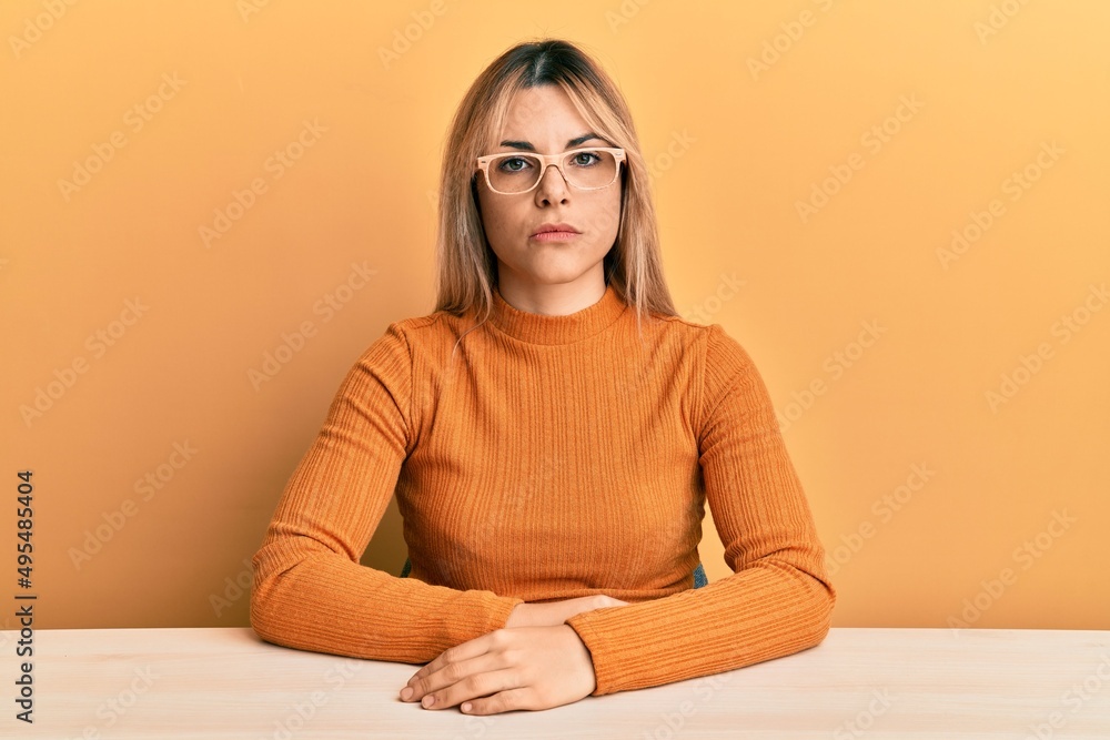 Young caucasian woman wearing casual clothes and glasses sitting on the table with serious expression on face. simple and natural looking at the camera. Young caucasian woman wearing casual clothes and glasses sitting on the table with serious expression on face. simple and natural looking at the camera.