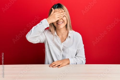 Young caucasian woman wearing casual clothes sitting on the table smiling and laughing with hand on face covering eyes for surprise. blind concept.