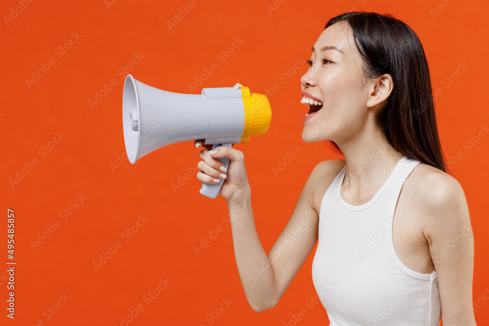 Side profile view jubilant young woman of Asian ethnicity 20s year old in white tank top hold scream in megaphone announces discounts sale Hurry up isolated on plain orange background studio portrait Side profile view jubilant young woman of Asian ethnicity 20s year old in white tank top hold scream in megaphone announces discounts sale Hurry up isolated on plain orange background studio portrait