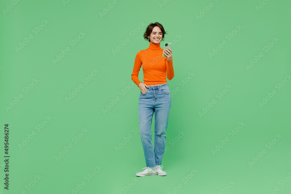 Full body young fun smiling woman 20s wear casual orange turtleneck hold in hand use mobile cell phone isolated on plain pastel light green color background studio portrait. People lifestyle concept. Full body young fun smiling woman 20s wear casual orange turtleneck hold in hand use mobile cell phone isolated on plain pastel light green color background studio portrait. People lifestyle concept.