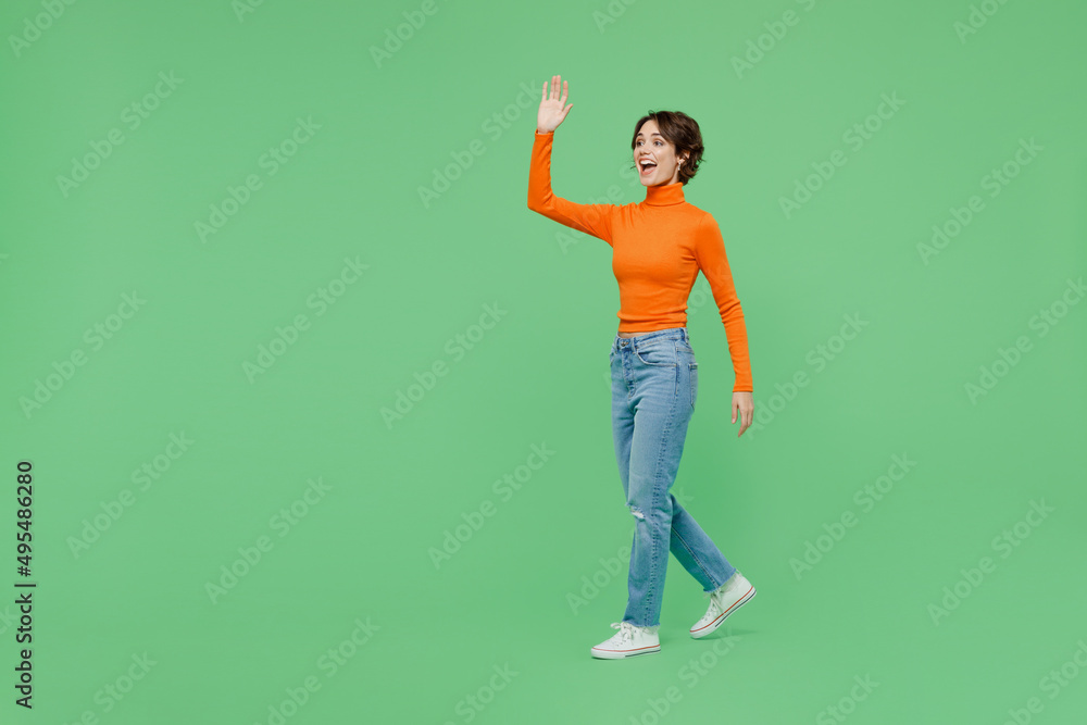 Full body young smiling happy cool fun woman 20s wear casual orange turtleneck walk going waving hand isolated on plain pastel light green color background studio portrait. People lifestyle concept. Full body young smiling happy cool fun woman 20s wear casual orange turtleneck walk going waving hand isolated on plain pastel light green color background studio portrait. People lifestyle concept.