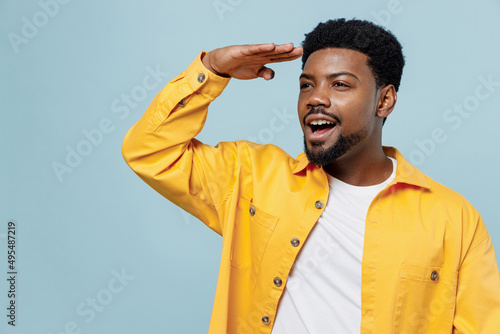 Young fun man of African American ethnicity 20s wear yellow shirt hold hand at forehead look far away distance isolated on plain pastel light blue background studio portrait. People lifestyle concept.