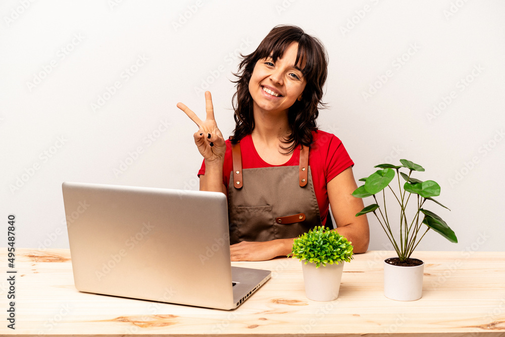 Young gardener hispanic woman sitting on a table isolated on white background joyful and carefree showing a peace symbol with fingers. Young gardener hispanic woman sitting on a table isolated on white background joyful and carefree showing a peace symbol with fingers.