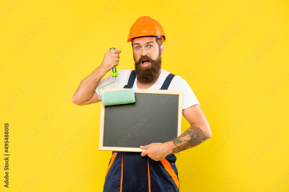 surprised man in helmet and work clothes hold paint roller and blackboard with copy space surprised man in helmet and work clothes hold paint roller and blackboard with copy space