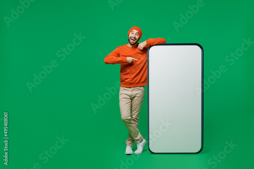 Full body young smiling fun man 20s wear orange sweatshirt hat point index finger on big blank screen mobile cell phone with workspace copy space mockup area isolated on plain green background studio.