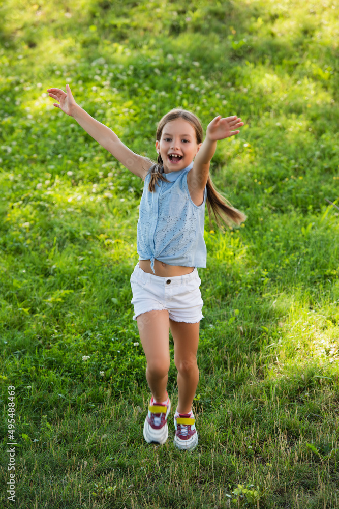 full length view of excited girl in shorts waving hands while running on green lawn. full length view of excited girl in shorts waving hands while running on green lawn.
