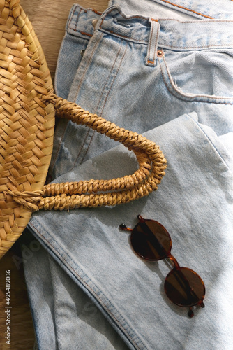 Vintage mom jeans, round straw bag and sunglasses on wooden background. Flat lay.