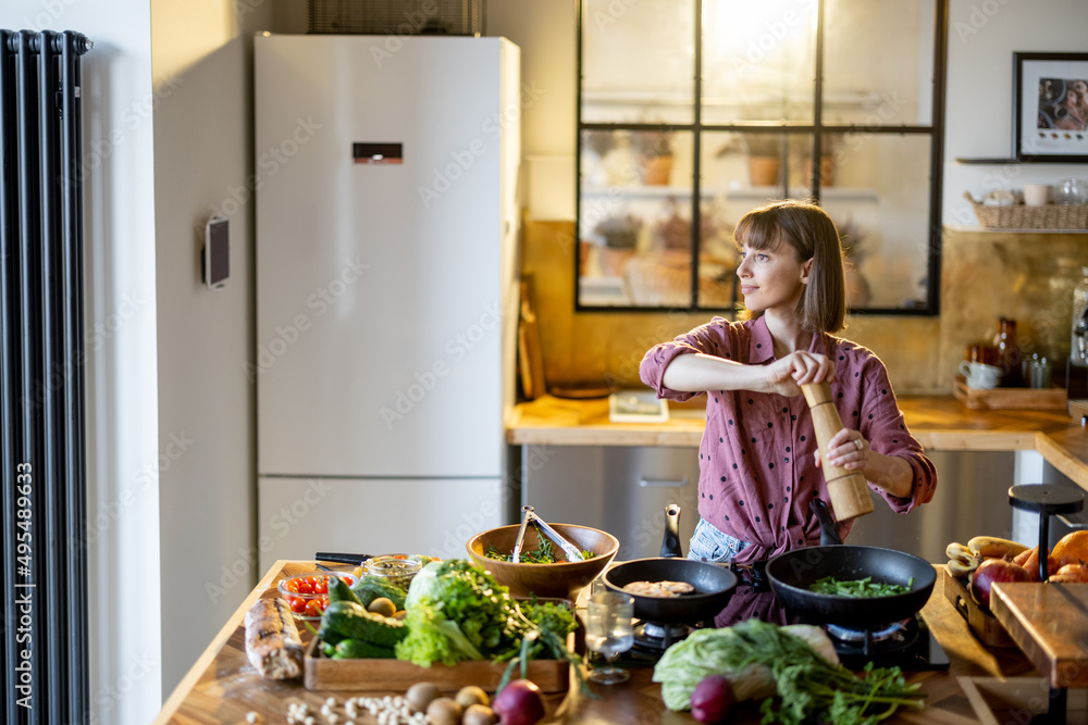 Young woman fries vegetables and seasons them on cooking pan while preparing healthy food at home. Lots of fresh food ingredients on table. Modern kitchen interior