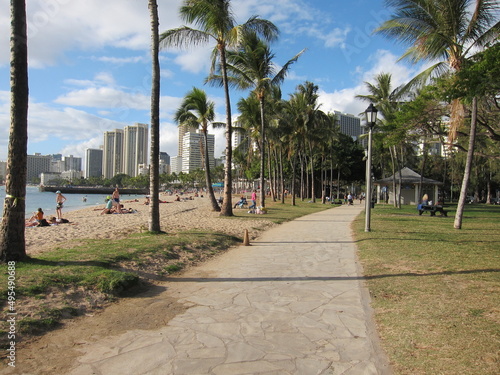 Walkway along the beach. View of green grass, lampposts, skyline, palm trees and blue sky with clouds. Oahu, Waikiki, Hawaii. People resting and strolling.