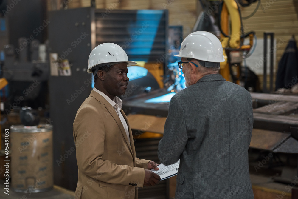 Rear view of two engineers in suit and hardhats discussing the work of machine in the factory Rear view of two engineers in suit and hardhats discussing the work of machine in the factory