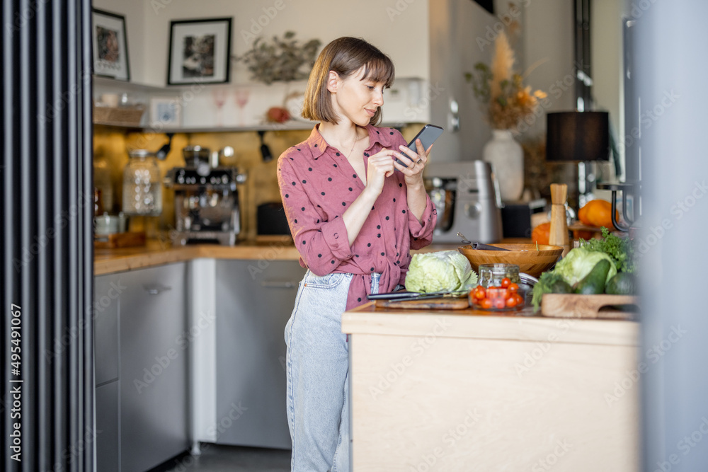 Young woman using smatphone while standing in the kitchen with lots of fresh green food ingredients on background. Housewife with phone at home