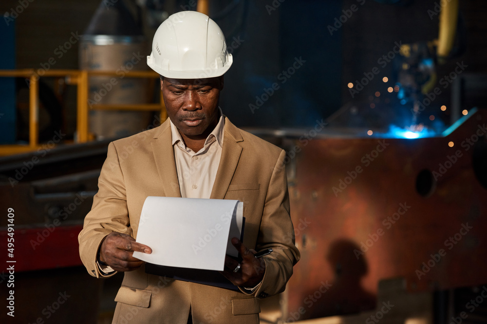 African serious engineer in hardhat examining the working plan on papers while standing in workshop in factory African serious engineer in hardhat examining the working plan on papers while standing in workshop in factory