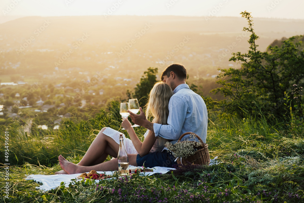 Beautiful couple having romantic picnic with tasty food and wine, sitting together on the picnic blanket at the hill on a sunset.