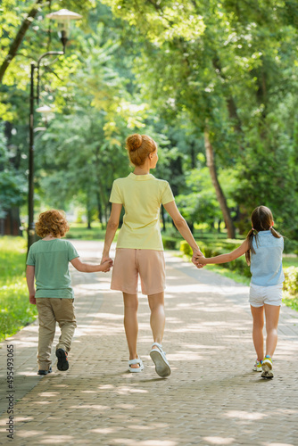 back view of woman with kids holding hands while walking in park.