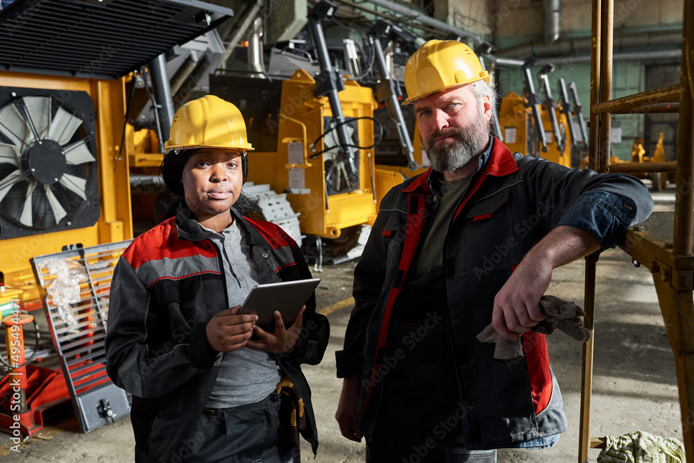Portrait of couple of technicians looking at camera while standing in warehouse, they controlling the machines with digital tablet Portrait of couple of technicians looking at camera while standing in warehouse, they controlling the machines with digital tablet