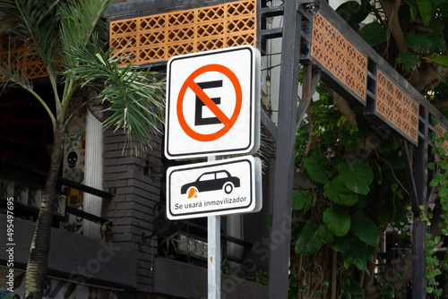 Road sign against the backdrop of dense foliage and old city. No parking sign. The inscription on the sign in Spanish - so will use immobilizer