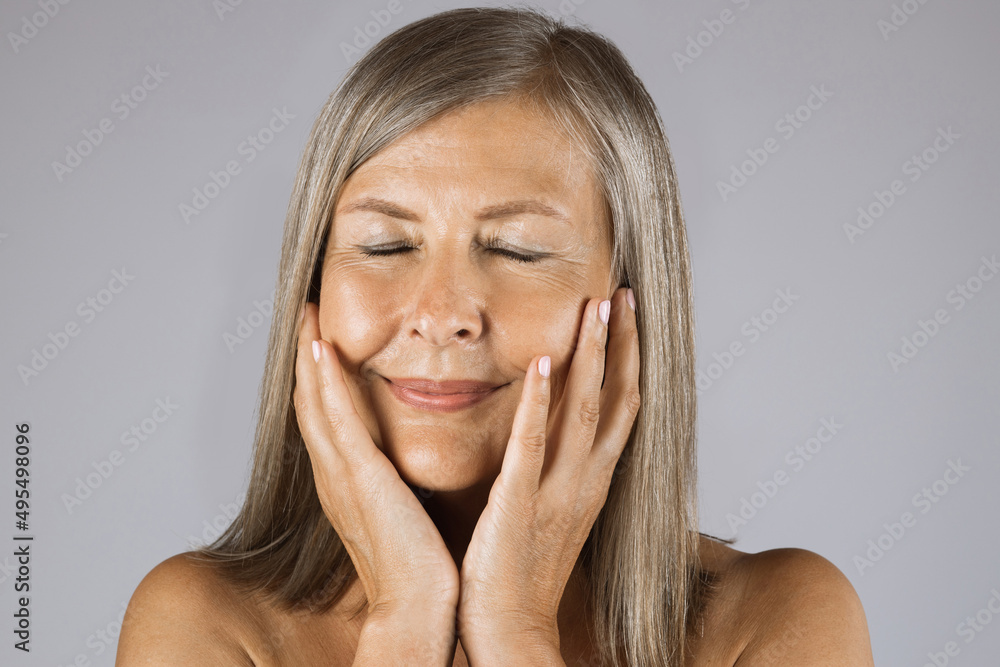 Portrait of pleasant aged woman keeping eyes closed and touching her cheeks. Isolated over grey studio background. Concept of skincare and beauty. Portrait of pleasant aged woman keeping eyes closed and touching her cheeks. Isolated over grey studio background. Concept of skincare and beauty.