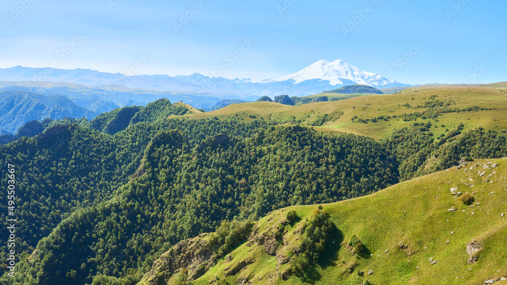 View of the sun-drenched green hills stretching in front of the peaks of Elbrus. Caucasus