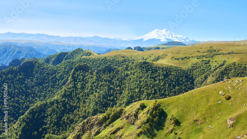 View of the sun-drenched green hills stretching in front of the peaks of Elbrus. Caucasus