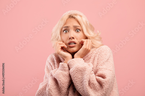 Young caucasian woman feeling scared, touching cheeks with hands, posing on pink studio background