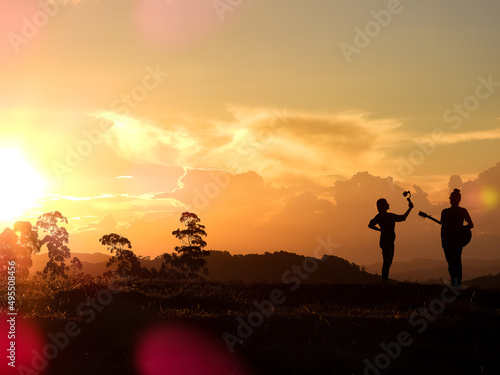 Fototapeta Naklejka Na Ścianę i Meble -  Orange sun setting in the mountains.  Man playing guitar silhouette and woman filming with gimbal and cell phone