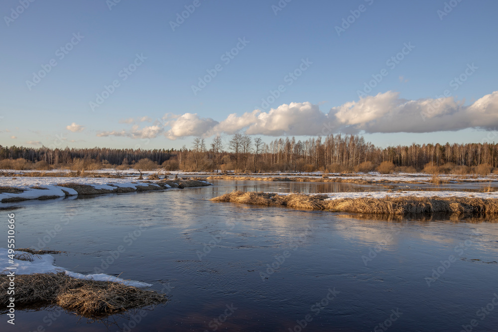 March sunny evening by the river. Blue sky over the horizon. A picturesque landscape, early spring, a river with snow-covered banks, dry grass and bushes. The first thaws, the snow is melting