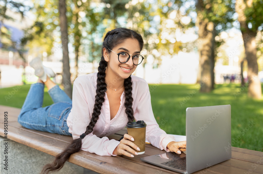 Happy modern student girl studying outdoors, resting with laptop and cup of coffee takeaway on bench in park Happy modern student girl studying outdoors, resting with laptop and cup of coffee takeaway on bench in park