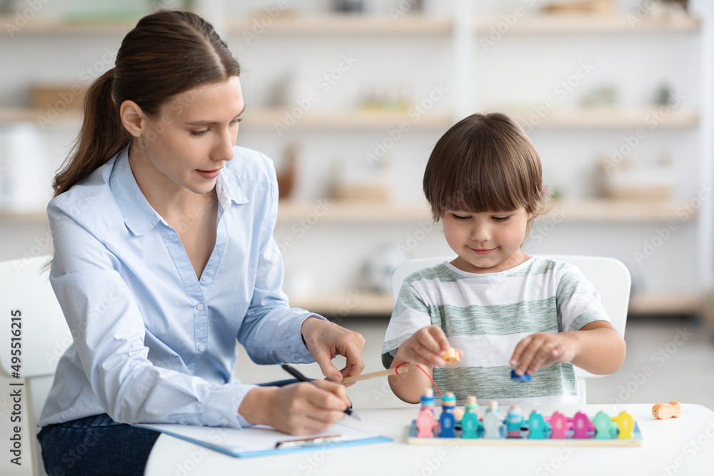Cute little boy playing colorful wooden game at specialist office, professional woman therapist taking notes, free space Cute little boy playing colorful wooden game at specialist office, professional woman therapist taking notes, free space