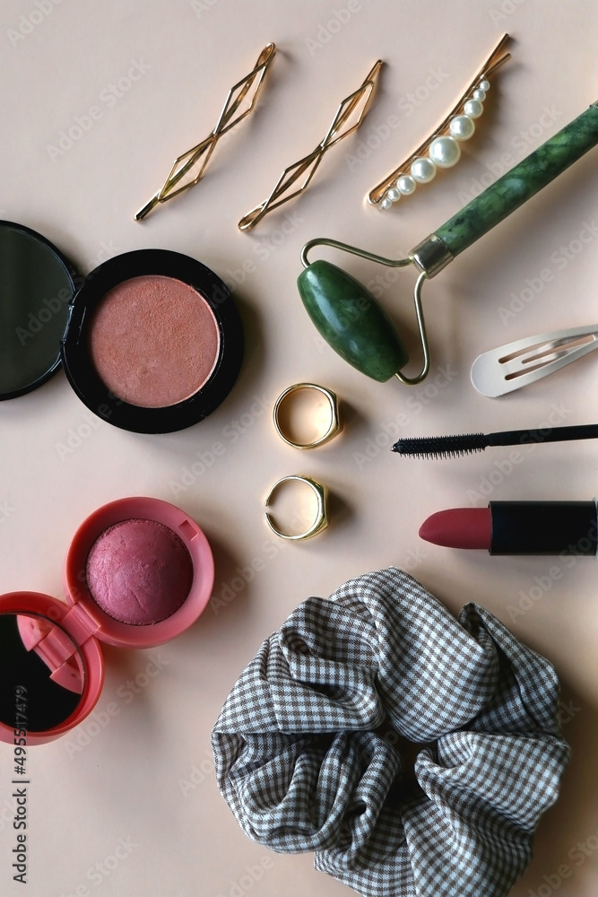 Gingham scrunchie, blush and highlighter, gold rings, various hair clips, jade roller, lipstick and mascara on beige background. Flat lay. Gingham scrunchie, blush and highlighter, gold rings, various hair clips, jade roller, lipstick and mascara on beige background. Flat lay.