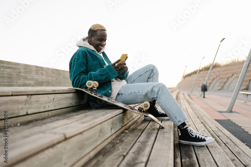 Happy young african man with skateboard looking at mobile phone social media app to connect with friends
