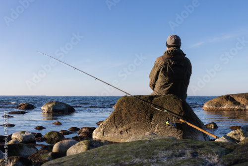 Meerforellen Angler mit Spinnrute und Küstenwobbler  sitzt auf Stein und blickt auf die Ostsee.