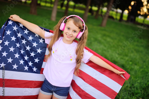 a little girl in denim shorts and pink headphones smiles and holds a large flag of the United States of America in her hands