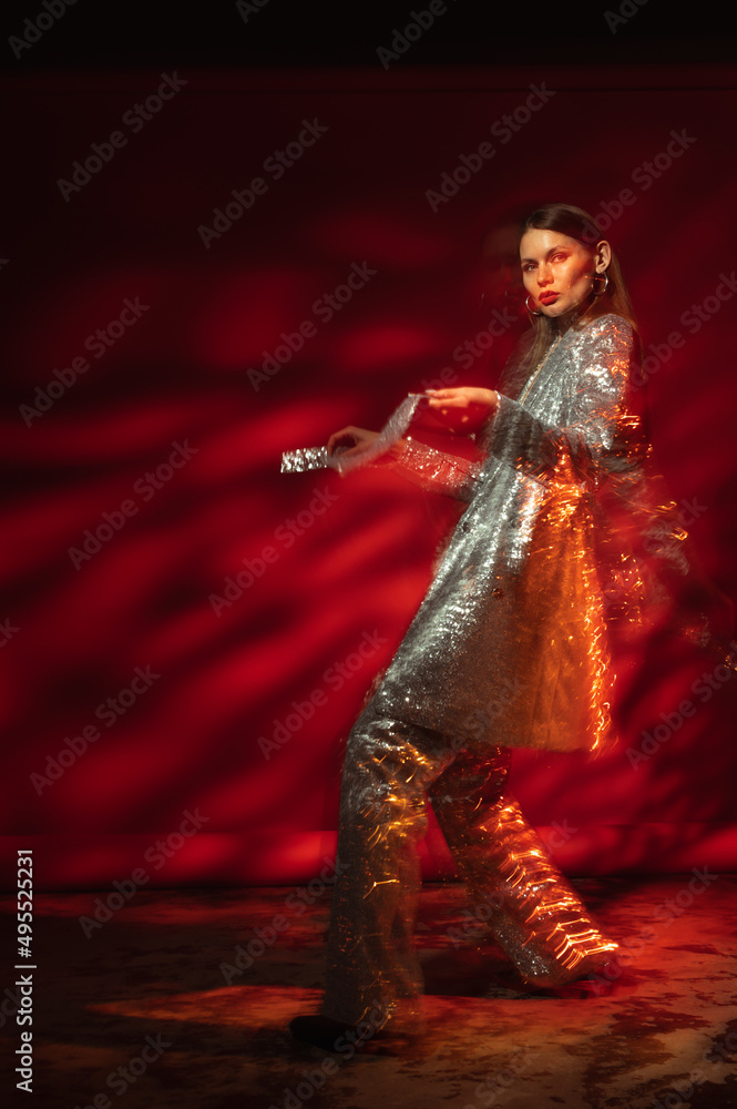 Fototapeta premium Fashionable woman wearing sequin suit, posing, dancing against red background. Full-length studio fashion portrait with move effect and art lighting