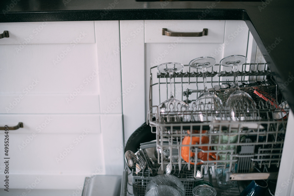 Open dishwasher with clean dishes in a white kitchen. Clean glasses, plates, mugs in the dishwasher basket Open dishwasher with clean dishes in a white kitchen. Clean glasses, plates, mugs in the dishwasher basket