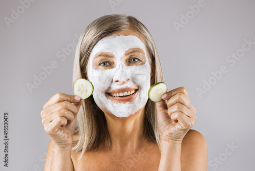 Portrait of positive caucasian woman holding slices of cucumber in hands while wearing white cosmetic mask on face. Isolated over grey studio background.