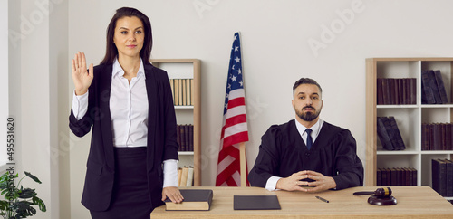 American witness takes an oath before giving a testimony. A jury member standing near a male judge in the court room swears on the Bible that she will tell the truth. Law and justice in the US concept photo