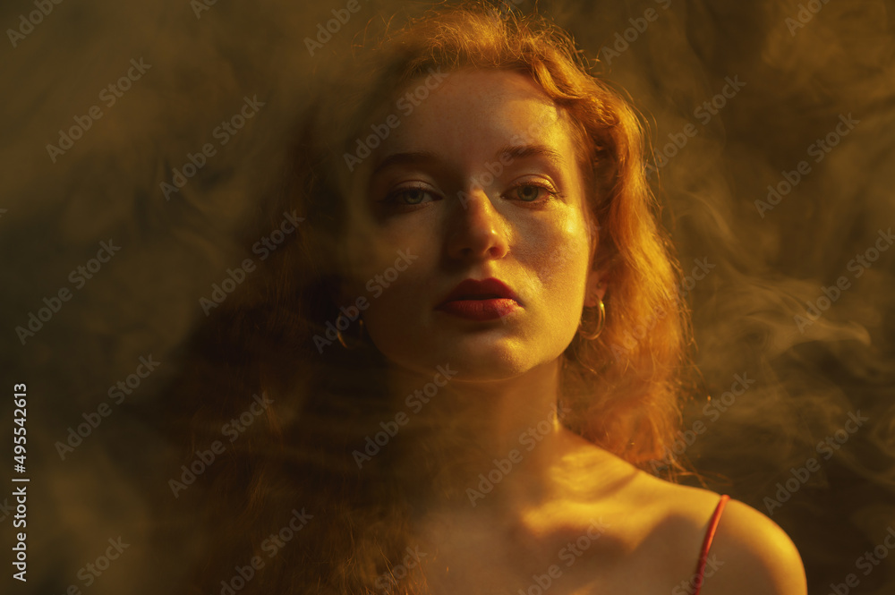 Portrait of beautiful redhead freckled woman posing in darkness, smoke and warm light