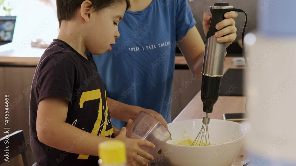 Mom and son standing near table in kitchen, boy adding milk into glass bowl cooking biscuits. Boy.add ingredients to bowl to make cookies. Beautiful with boy Mom and son standing near table in kitchen, boy adding milk into glass bowl cooking biscuits. Boy.add ingredients to bowl to make cookies. Beautiful with boy