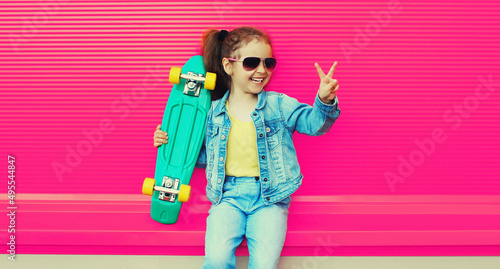 Portrait of cheerful laughing little girl child posing with skateboard in the city on colorful background
