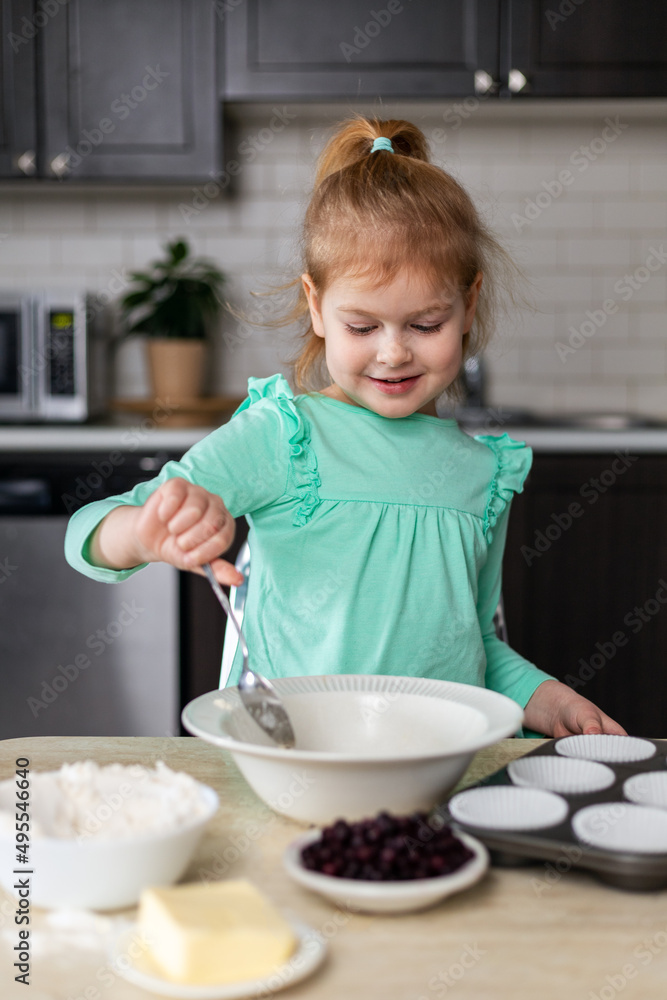 Little smiling girl making dough in kitchen. Small happy child cooking. Kid making homemade muffins Little smiling girl making dough in kitchen. Small happy child cooking. Kid making homemade muffins