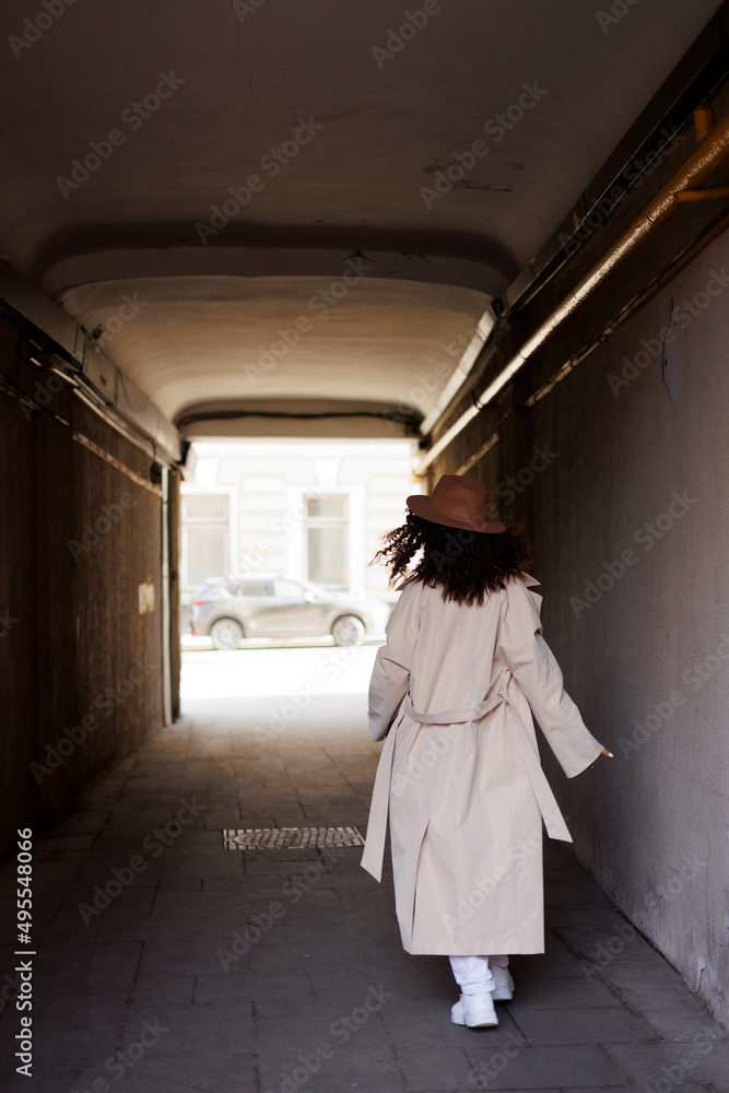  curly brunette girl in sun glasses and hat going away outdoors. Young woman happy walking in street. Pink sweater, beige coat, beige hat. Sun in city. Fashionable asian girl with frizzly hair