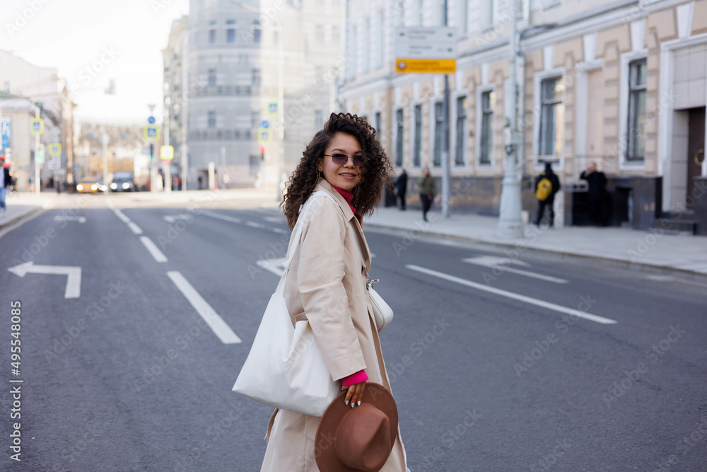 Happy curly brunette girl in sun glasses smiling outdoors. Young woman happy walking in street. Pink sweater, beige coat, beige hat. Sun in city. Fashionable asian girl with frizzly hair
