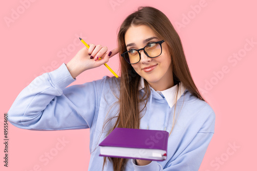 Jiyful student girl with long blond hair in glasses holding a book in one hand and keeping a pencil in another holding an index finget on the temple.