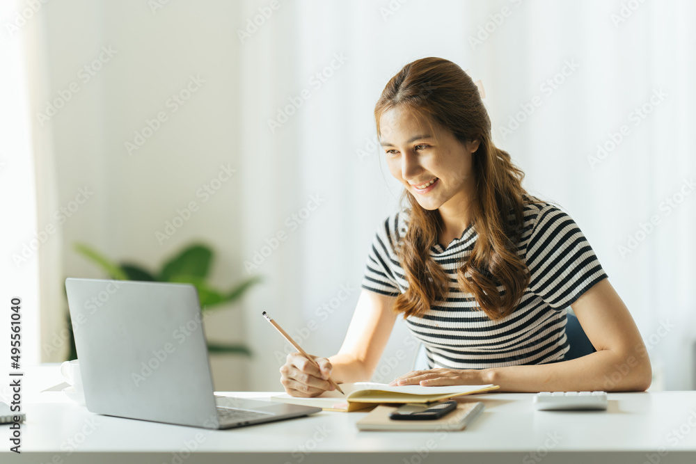 Young women take notes in their home office while working on a laptop. Young women take notes in their home office while working on a laptop.