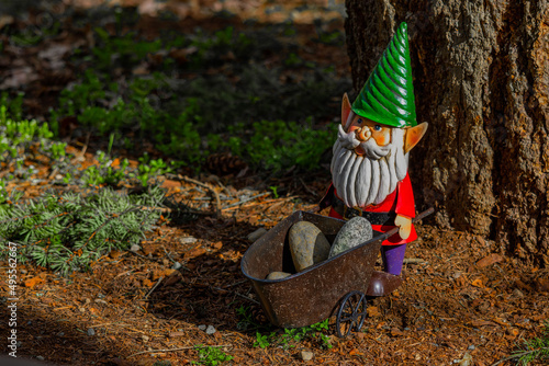 Metal lawn gnome holding pile of rocks in wheelbarrow standing on top of beauty bark and debris.