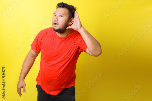 Young attractive Asian fat man serously listening, posing hand over ear listening an hearing to rumor or gossip. Deafness concept.