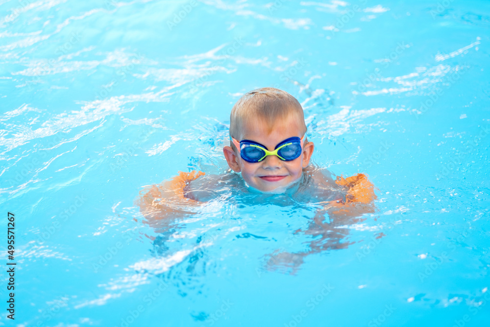 Naklejka premium Portrait smiling boy in swimming pool, child in swimming glasses and inflatable sleeves. Summer travel hotel vacation or classes