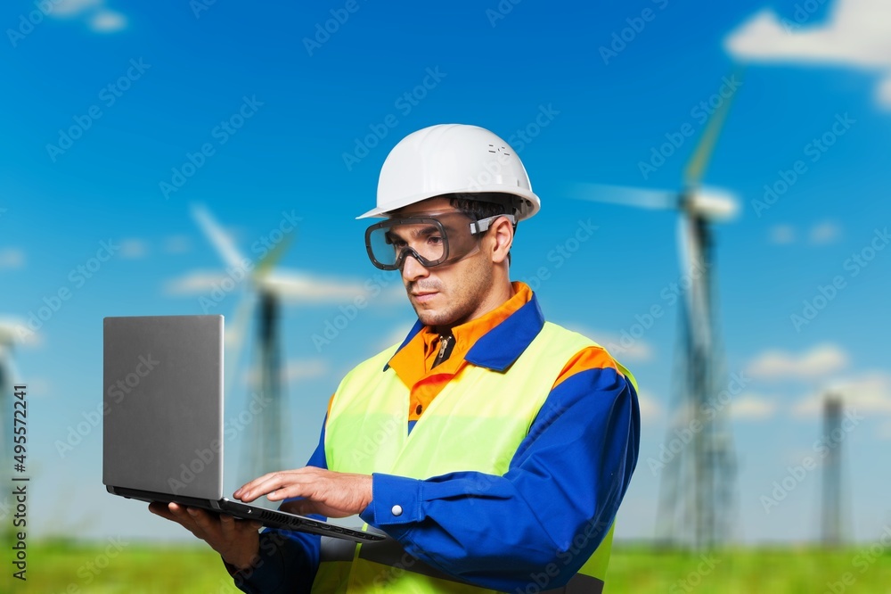 Beautiful man in white helmet working with digital tablet at energy farm. Male inspector controlling functioning of wind turbines outdoors. Beautiful man in white helmet working with digital tablet at energy farm. Male inspector controlling functioning of wind turbines outdoors.