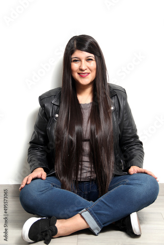 Happy and proud Latin young adult woman shows her long straight, black and shiny hair on a white background with a leather jacket and black tennis shoes 