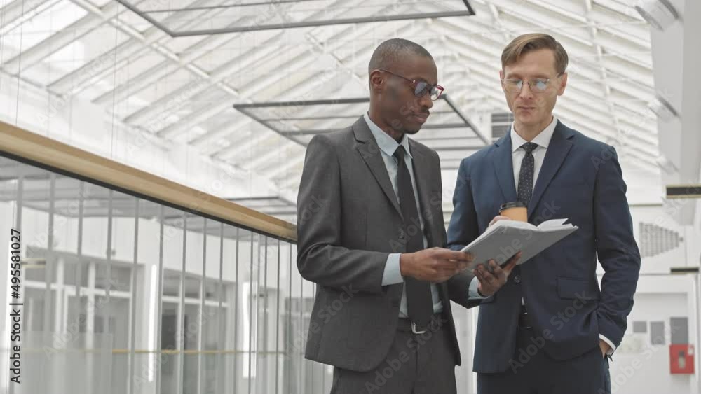 Medium slowmo of two successful multiethnic businessmen standing at modern business center with notebook discussing common project then shaking hands in agreement Medium slowmo of two successful multiethnic businessmen standing at modern business center with notebook discussing common project then shaking hands in agreement